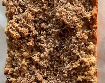 May include: A loaf of baked bread with a crumb topping. The bread is light brown with a textured, crumbly topping. The bread is rectangular and sits on a white surface.
