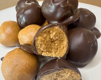 May include: A selection of peanut butter balls, some covered in dark chocolate. One is cut open, showing the peanut butter centre. The treats are presented on a white plate, against a light brown backdrop.