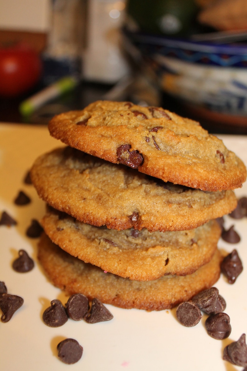 K&ouml;nnte beinhalten: Ein Stapel von vier goldbraunen Schokoladen-Chip-Cookies. Jeder Keks ist rund und hat sichtbare Schokoladenst&uuml;ckchen. Einige Schokoladenst&uuml;ckchen sind um die Basis des Stapels verstreut. Die Kekse sehen frisch gebacken aus.