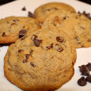 K&ouml;nnte beinhalten: Nahaufnahme von mehreren frisch gebackenen Schokoladen-Chip-Cookies auf einem wei&szlig;en Teller. Die Kekse sind goldbraun mit sichtbaren Schokoladenst&uuml;ckchen. Einige Schokoladenst&uuml;ckchen sind verstreut.