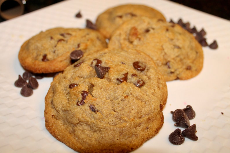 K&ouml;nnte beinhalten: Nahaufnahme von mehreren frisch gebackenen Schokoladen-Chip-Cookies auf einem wei&szlig;en Teller. Die Kekse sind goldbraun mit sichtbaren Schokoladenst&uuml;ckchen. Einige Schokoladenst&uuml;ckchen sind verstreut.