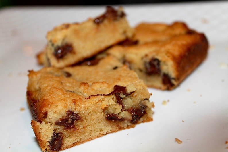 Puede incluir: Primer plano de varias barras de galletas con chispas de chocolate doradas sobre un plato blanco. Las barras tienen una textura suave y similar a la de un pastel con chispas de chocolate visibles. La imagen muestra los productos horneados.