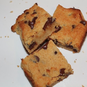 Puede incluir: Tres barras de galletas con chispas de chocolate cortadas en cuadrados sobre una superficie blanca. Las barras tienen un exterior marr&oacute;n dorado y est&aacute;n salpicadas de chispas de chocolate negro. Parte del chocolate se ha derretido, creando una textura pegajosa.
