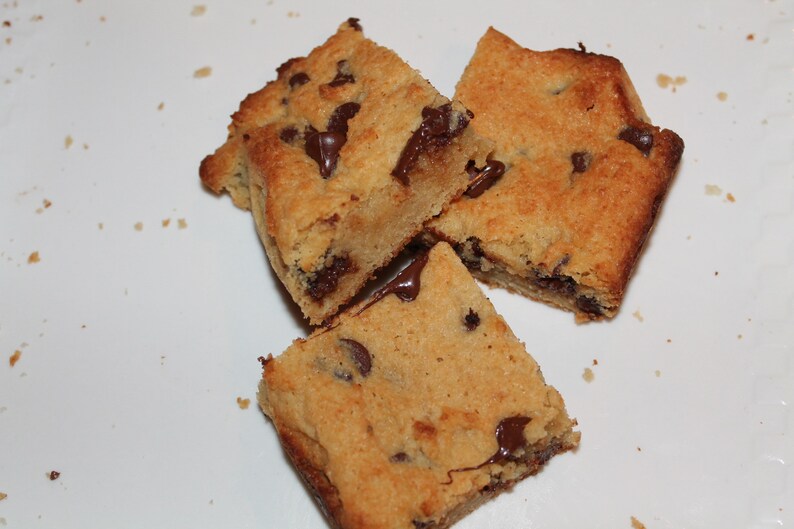 Puede incluir: Tres barras de galletas con chispas de chocolate cortadas en cuadrados sobre una superficie blanca. Las barras tienen un exterior marr&oacute;n dorado y est&aacute;n salpicadas de chispas de chocolate negro. Parte del chocolate se ha derretido, creando una textura pegajosa.
