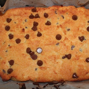 Puede incluir: Una barra de galletas con chispas de chocolate reci&eacute;n horneada, de color marr&oacute;n dorado, salpicada de chispas de chocolate negro. La barra de galletas est&aacute; horneada en una fuente para hornear forrada con papel pergamino, con una superficie ligeramente irregular y bordes crujientes.
