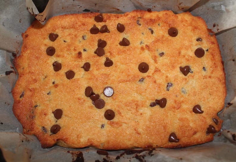 Puede incluir: Una barra de galletas con chispas de chocolate reci&eacute;n horneada, de color marr&oacute;n dorado, salpicada de chispas de chocolate negro. La barra de galletas est&aacute; horneada en una fuente para hornear forrada con papel pergamino, con una superficie ligeramente irregular y bordes crujientes.