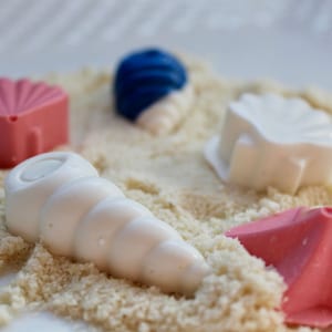 May include: An assortment of seashell-shaped chocolates in white, pink, and blue, arranged on a bed of sand-like crumbs. The chocolates include a conch shell, a starfish, and other geometric shapes.