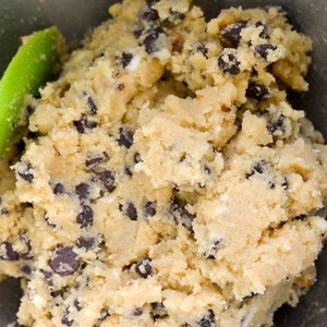 K&ouml;nnte beinhalten: Nahaufnahme von Keksteig in einer Edelstahlsch&uuml;ssel. Der Teig ist hellbeige und mit dunklen Schokoladenst&uuml;ckchen gesprenkelt. Auf der linken Seite ist ein gr&uuml;ner Spatel zu sehen, was darauf hindeutet, dass der Teig verr&uuml;hrt wird.