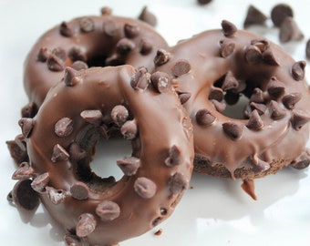 May include: Three chocolate doughnuts with a dark brown glaze and scattered chocolate chips. The doughnuts are arranged on a white surface, with extra chips around them. A sweet treat.