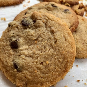 K&ouml;nnte beinhalten: Nahaufnahme von mehreren frisch gebackenen Schokoladen-Chip-Cookies. Die Kekse sind goldbraun mit sichtbaren Schokoladenst&uuml;ckchen und einer leicht strukturierten Oberfl&auml;che. Einige Keksbr&ouml;sel sind um die Kekse verstreut.