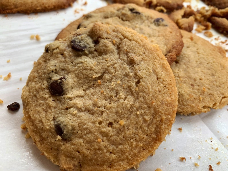 K&ouml;nnte beinhalten: Nahaufnahme von mehreren frisch gebackenen Schokoladen-Chip-Cookies. Die Kekse sind goldbraun mit sichtbaren Schokoladenst&uuml;ckchen und einer leicht strukturierten Oberfl&auml;che. Einige Keksbr&ouml;sel sind um die Kekse verstreut.