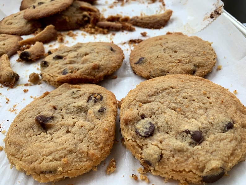 K&ouml;nnte beinhalten: Nahaufnahme von mehreren frisch gebackenen Schokoladen-Chip-Cookies auf einem Backblech. Die Kekse sind goldbraun mit sichtbaren Schokoladenst&uuml;ckchen. Einige Kekse sind ganz, andere haben zerbrochene St&uuml;cke.