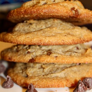 K&ouml;nnte beinhalten: Ein Stapel von vier goldbraunen Schokoladen-Chip-Cookies. Die Kekse haben eine leicht strukturierte Oberfl&auml;che und sichtbare Schokoladenst&uuml;ckchen. Einige Schokoladenst&uuml;ckchen sind um die Basis des Stapels verstreut.