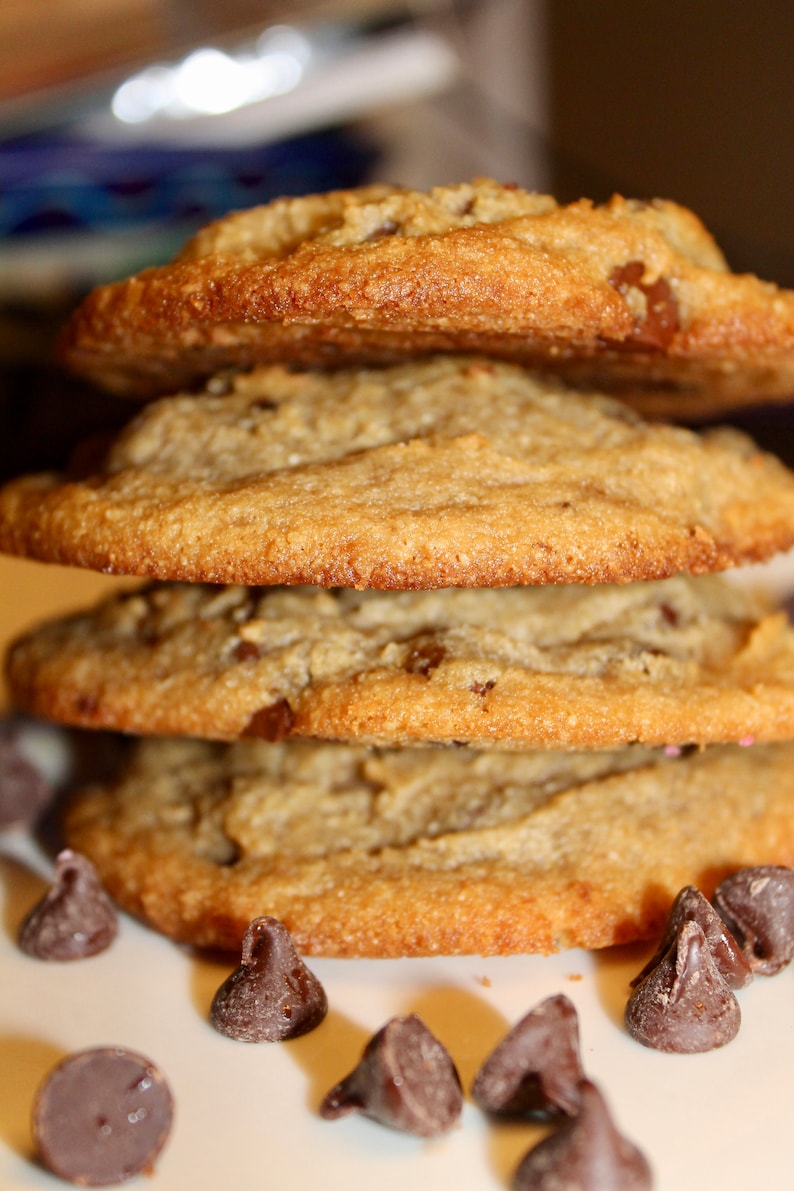 K&ouml;nnte beinhalten: Ein Stapel von vier goldbraunen Schokoladen-Chip-Cookies. Die Kekse haben eine leicht strukturierte Oberfl&auml;che und sichtbare Schokoladenst&uuml;ckchen. Einige Schokoladenst&uuml;ckchen sind um die Basis des Stapels verstreut.