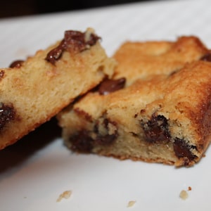 Puede incluir: Primer plano de varias barras de galletas de color marr&oacute;n dorado con chispas de chocolate visibles. Las barras est&aacute;n cortadas en formas rectangulares y parecen reci&eacute;n horneadas, con un exterior ligeramente crujiente y un interior suave. La imagen est&aacute; sobre una superficie blanca.