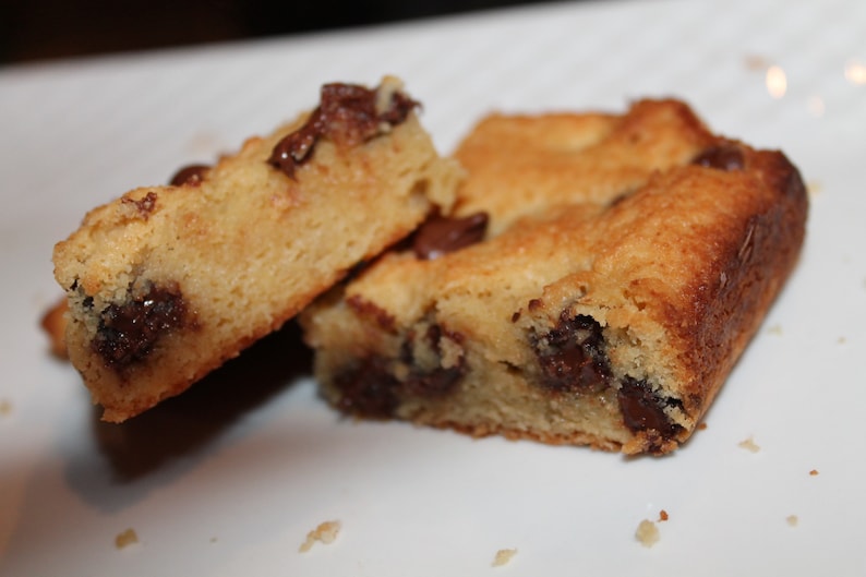 Puede incluir: Primer plano de varias barras de galletas de color marr&oacute;n dorado con chispas de chocolate visibles. Las barras est&aacute;n cortadas en formas rectangulares y parecen reci&eacute;n horneadas, con un exterior ligeramente crujiente y un interior suave. La imagen est&aacute; sobre una superficie blanca.