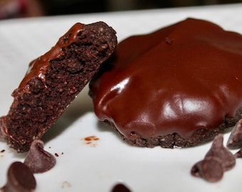 Puede incluir: Primer plano de un postre de chocolate sobre un plato blanco. Una pieza está cortada por la mitad, revelando el interior oscuro. La otra está entera, cubierta con una capa brillante de chocolate. Hay chispas de chocolate esparcidas.