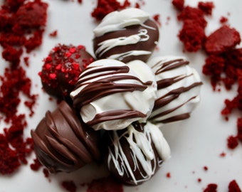 May include: A close-up of several chocolate-covered cake pops. Some are drizzled with white chocolate, others with dark chocolate. One is covered in red heart-shaped sprinkles. Red velvet cake crumbs are scattered around the pops.