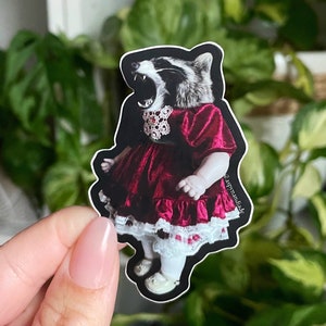 May include: A sticker of a raccoon wearing a red velvet dress with a white lace trim and a jeweled collar. The raccoon is looking up and has its mouth open, as if it is singing. The sticker is on a white background.