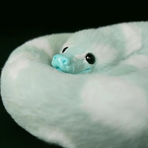 May include: A plush snake toy coiled in a circle, featuring soft mint green and white faux fur. The head is made of a smooth, light blue material with a textured scale pattern, featuring large, glossy black eyes and a small, rounded snout.