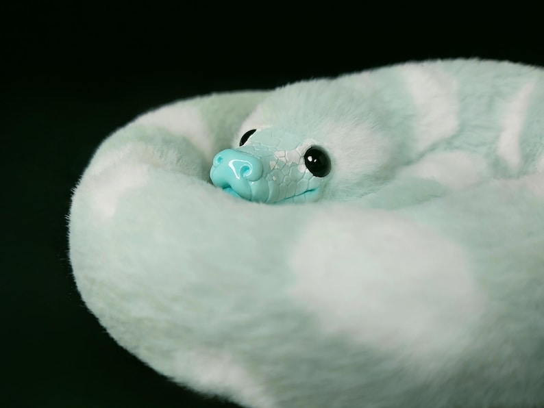 May include: A plush snake toy coiled in a circle, featuring soft mint green and white faux fur. The head is made of a smooth, light blue material with a textured scale pattern, featuring large, glossy black eyes and a small, rounded snout.