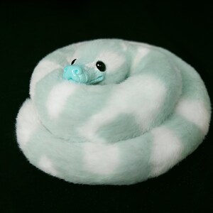 May include: A plush snake coiled in a circular shape, featuring soft faux fur with a pattern of alternating mint green and white patches. The toy has a smooth, bright turquoise plastic snout and large, glossy black eyes set into the head.