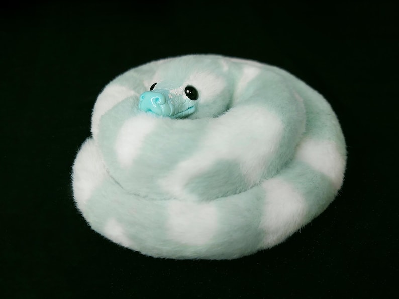 May include: A plush snake coiled in a circular shape, featuring soft faux fur with a pattern of alternating mint green and white patches. The toy has a smooth, bright turquoise plastic snout and large, glossy black eyes set into the head.