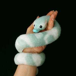 May include: A plush snake toy featuring a soft, fuzzy texture with a pattern of alternating light mint green and white patches. The snake has a smooth, solid light blue snout and two round, glossy black eyes positioned on the sides of its head.