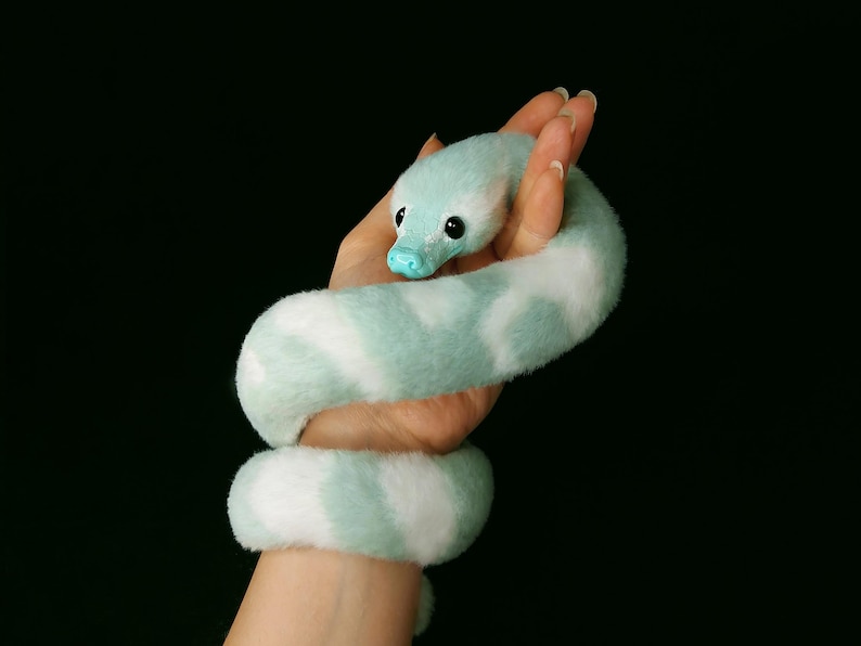 May include: A plush snake toy featuring a soft, fuzzy texture with a pattern of alternating light mint green and white patches. The snake has a smooth, solid light blue snout and two round, glossy black eyes positioned on the sides of its head.