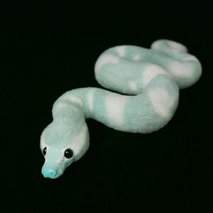 May include: A plush snake toy featuring a soft, fuzzy texture with a pattern of alternating light mint green and white segments. The snake has a smooth, solid mint green snout and two small, round, black eyes set against a black background.