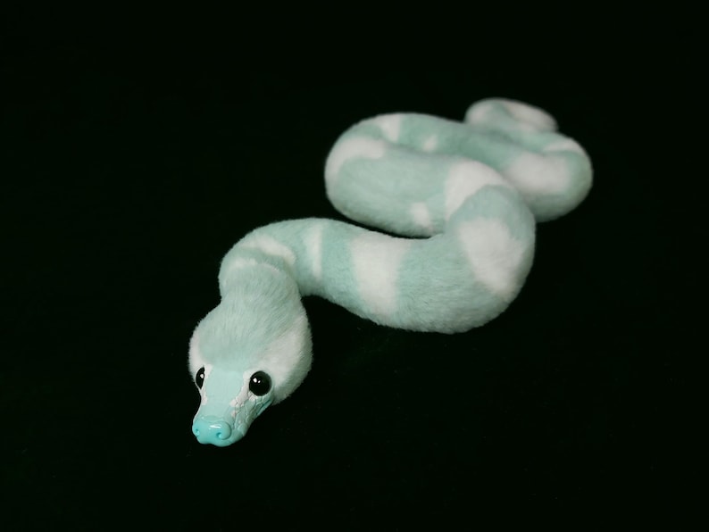 May include: A plush snake toy featuring a soft, fuzzy texture with a pattern of alternating light mint green and white segments. The snake has a smooth, solid mint green snout and two small, round, black eyes set against a black background.