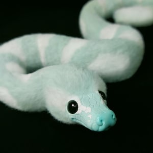 May include: A plush snake toy featuring a soft, fuzzy body with alternating light mint green and white stripes. The head is made of a smooth, solid light blue material with a textured scale pattern, large black eyes, and a defined snout.