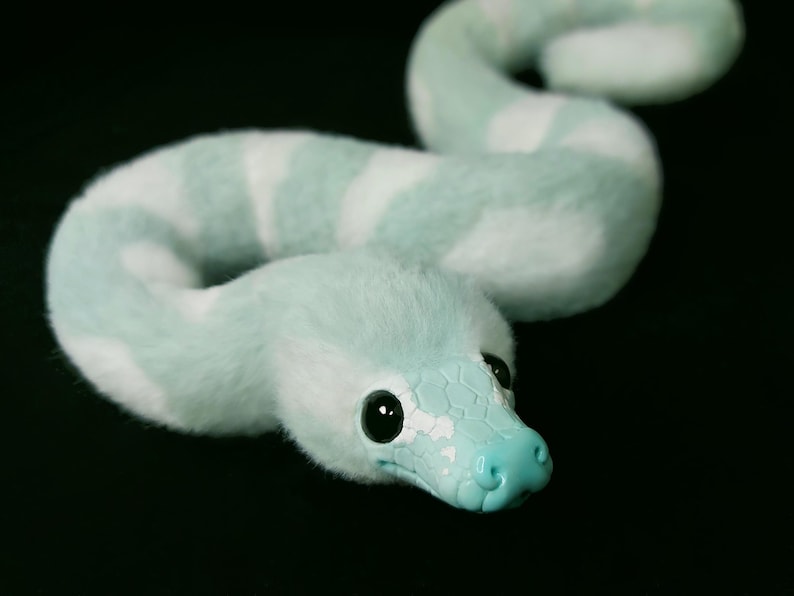 May include: A plush snake toy featuring a soft, fuzzy body with alternating light mint green and white stripes. The head is made of a smooth, solid light blue material with a textured scale pattern, large black eyes, and a defined snout.