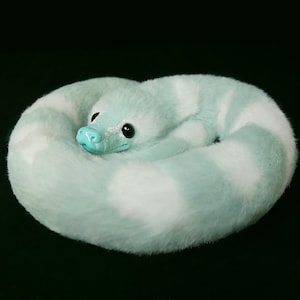 May include: A plush snake toy coiled in a circle, featuring alternating bands of soft mint green and white faux fur. The creature has a smooth, glossy mint green snout and large, dark, round eyes, creating a realistic yet soft appearance.