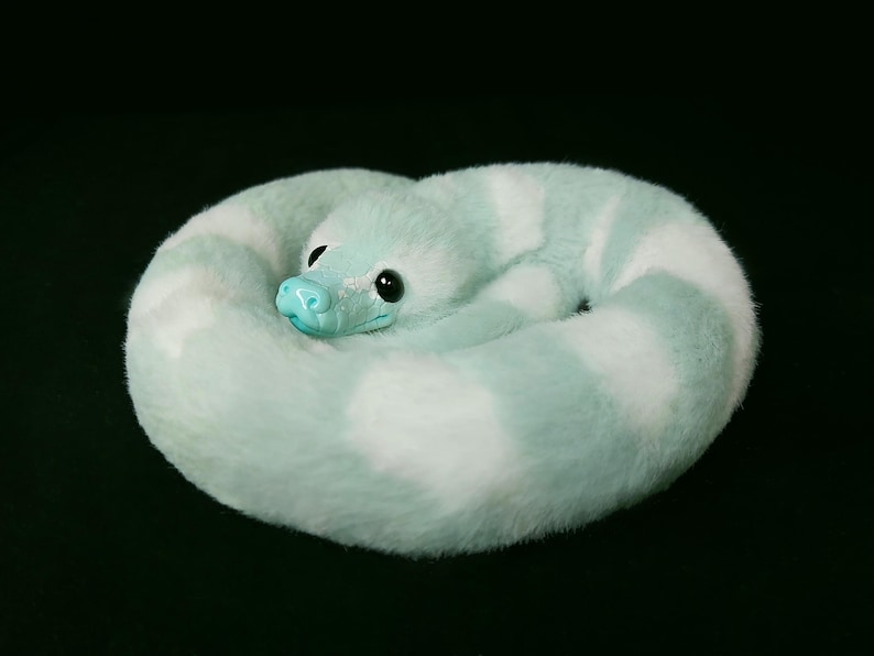 May include: A plush snake toy coiled in a circle, featuring alternating bands of soft mint green and white faux fur. The creature has a smooth, glossy mint green snout and large, dark, round eyes, creating a realistic yet soft appearance.