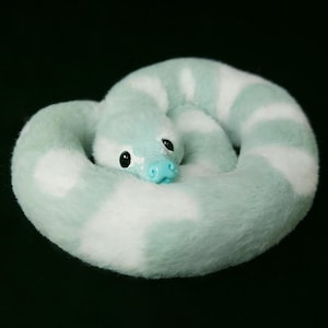 May include: A plush snake toy coiled in a circle, featuring a soft, fuzzy texture with alternating bands of light mint green and white. The snake has a smooth, bright turquoise plastic nose and large, round, glossy black eyes.