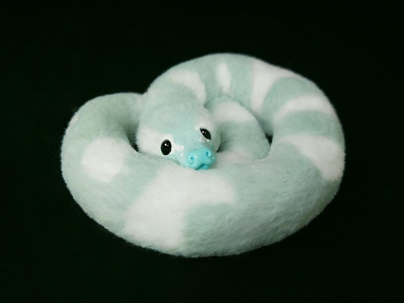 May include: A plush snake toy coiled in a circle, featuring a soft, fuzzy texture with alternating bands of light mint green and white. The snake has a smooth, bright turquoise plastic nose and large, round, glossy black eyes.