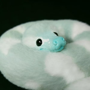 May include: A plush snake toy featuring a soft, fuzzy texture with a pattern of light mint green and white patches. The snake has a smooth, glossy light blue head with large, round black eyes and a sculpted snout, coiled into a circular shape.