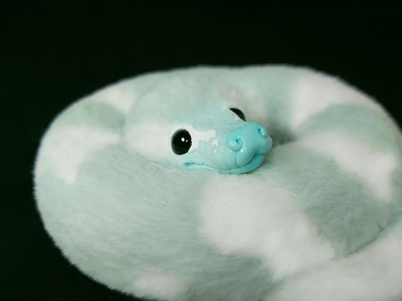 May include: A plush snake toy featuring a soft, fuzzy texture with a pattern of light mint green and white patches. The snake has a smooth, glossy light blue head with large, round black eyes and a sculpted snout, coiled into a circular shape.