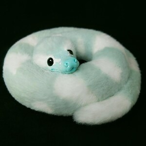 May include: A plush snake toy coiled in a circle, featuring soft faux fur with alternating patches of light mint green and white. The snake has a smooth, light blue plastic snout and large, glossy black eyes, creating a rounded, compact form.
