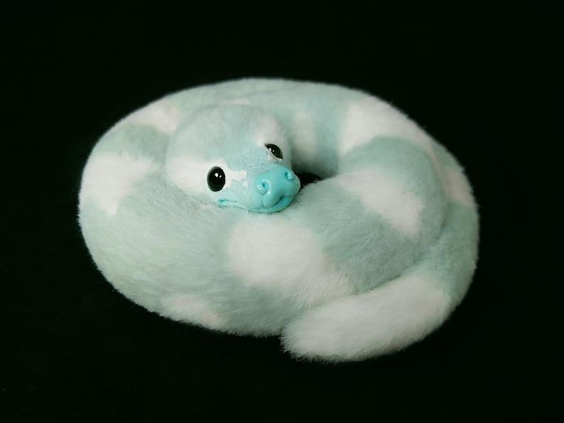 May include: A plush snake toy coiled in a circle, featuring soft faux fur with alternating patches of light mint green and white. The snake has a smooth, light blue plastic snout and large, glossy black eyes, creating a rounded, compact form.