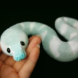May include: A plush snake toy featuring a soft, fuzzy body with alternating light mint green and white stripes. The head has a smooth, light blue plastic snout with a textured scale pattern, two black glossy eyes, and a curved, elongated body shape.