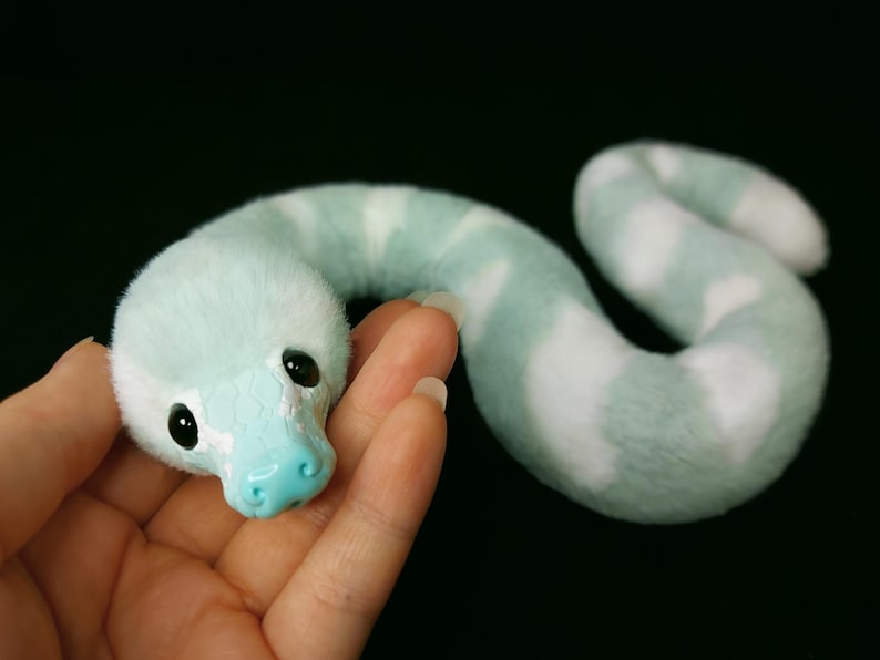 May include: A plush snake toy featuring a soft, fuzzy body with alternating light mint green and white stripes. The head has a smooth, light blue plastic snout with a textured scale pattern, two black glossy eyes, and a curved, elongated body shape.