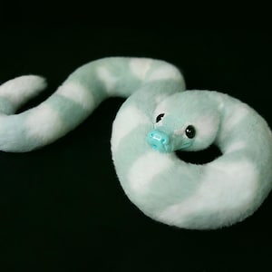May include: A plush snake toy featuring a soft, fuzzy texture with alternating bands of light mint green and white. The creature has a smooth, glossy light blue snout and large, dark, round eyes, coiled into a circular shape on a black background.