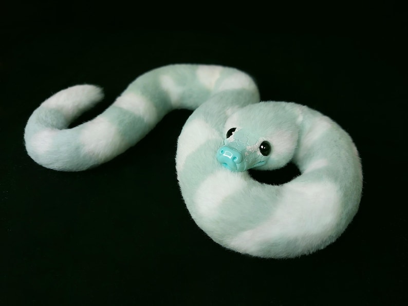 May include: A plush snake toy featuring a soft, fuzzy texture with alternating bands of light mint green and white. The creature has a smooth, glossy light blue snout and large, dark, round eyes, coiled into a circular shape on a black background.