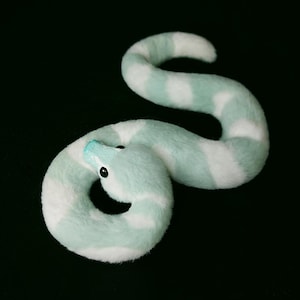 May include: A plush snake toy featuring a soft, fuzzy texture with alternating bands of mint green and white. The snake has a smooth, light blue snout and small, round, black eyes, coiled into a curved, serpentine shape against a dark background.