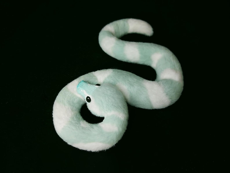 May include: A plush snake toy featuring a soft, fuzzy texture with alternating bands of mint green and white. The snake has a smooth, light blue snout and small, round, black eyes, coiled into a curved, serpentine shape against a dark background.