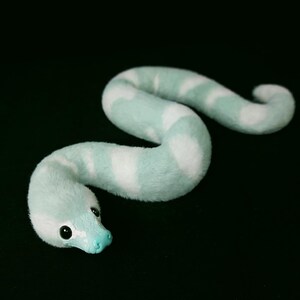 May include: A plush snake toy featuring a soft, fuzzy texture with alternating bands of light mint green and white. The snake has a smooth, solid mint green snout and two small, round, black eyes positioned on the sides of its head.