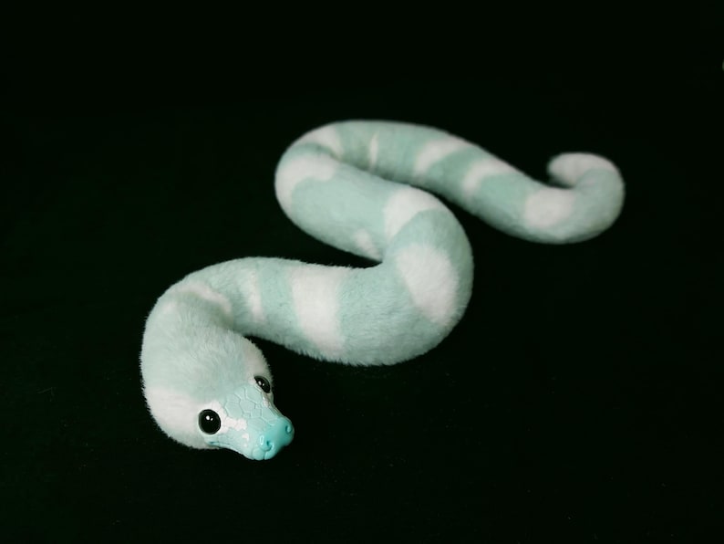 May include: A plush snake toy featuring a soft, fuzzy texture with alternating bands of light mint green and white. The snake has a smooth, solid mint green snout and two small, round, black eyes positioned on the sides of its head.