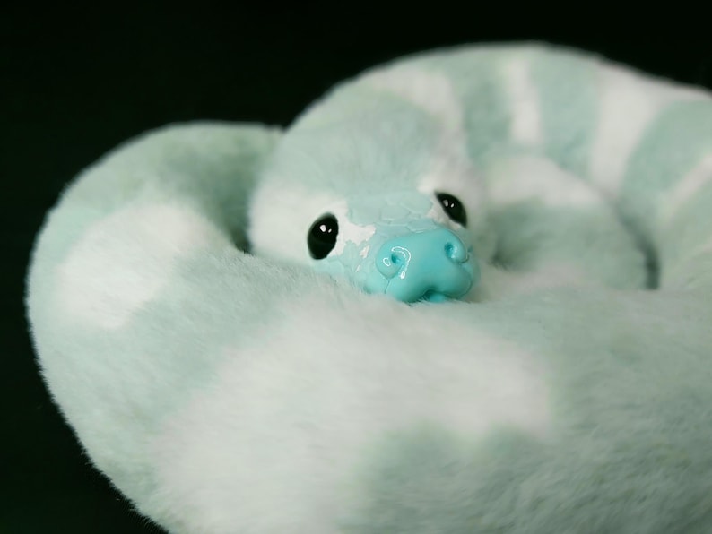 May include: A plush snake toy featuring soft, faux fur in alternating bands of mint green and white. The creature has a smooth, glossy, bright turquoise snout and two large, round, black eyes positioned centrally on its coiled, fuzzy body.